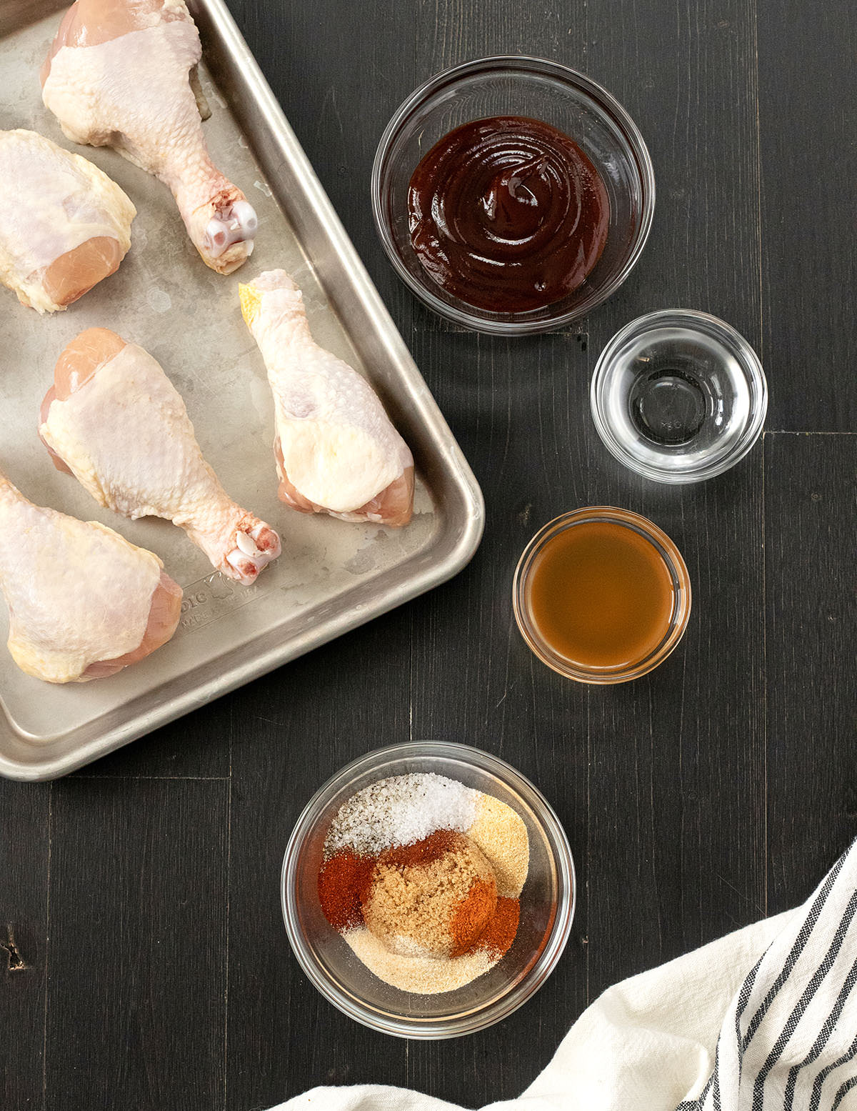 BBQ spices, bbq sauce and raw poultry legs on a baking sheet.