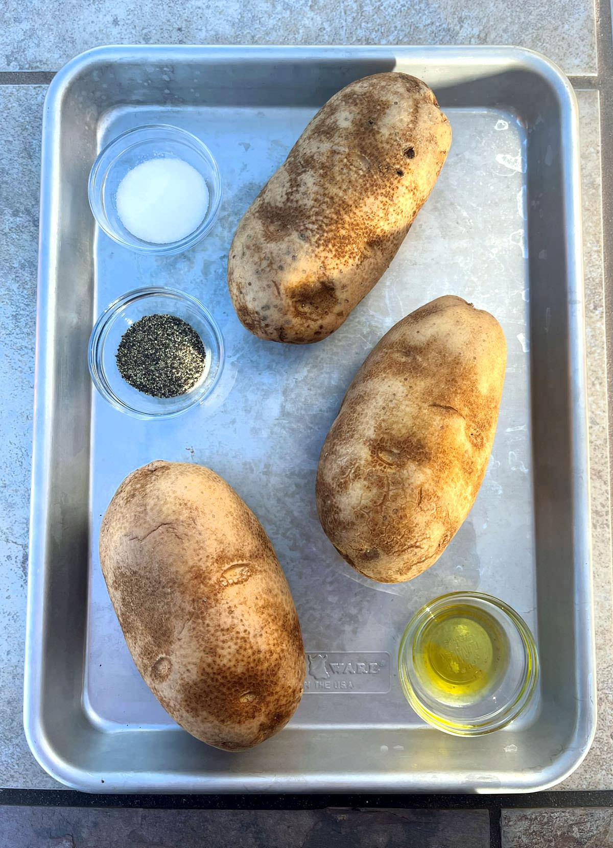 3 baked potatoes, salt and pepper on a silver baking sheet.